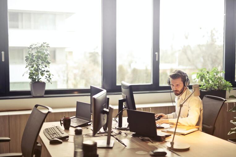 Hombre de barba trabajando al lado de un ventanal sobre mesa de trabajo con sus ordenadores y con auriculares.